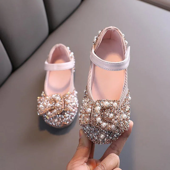 Pink baby shoes with pearl and rhinestone bow embellishments on a gray sofa