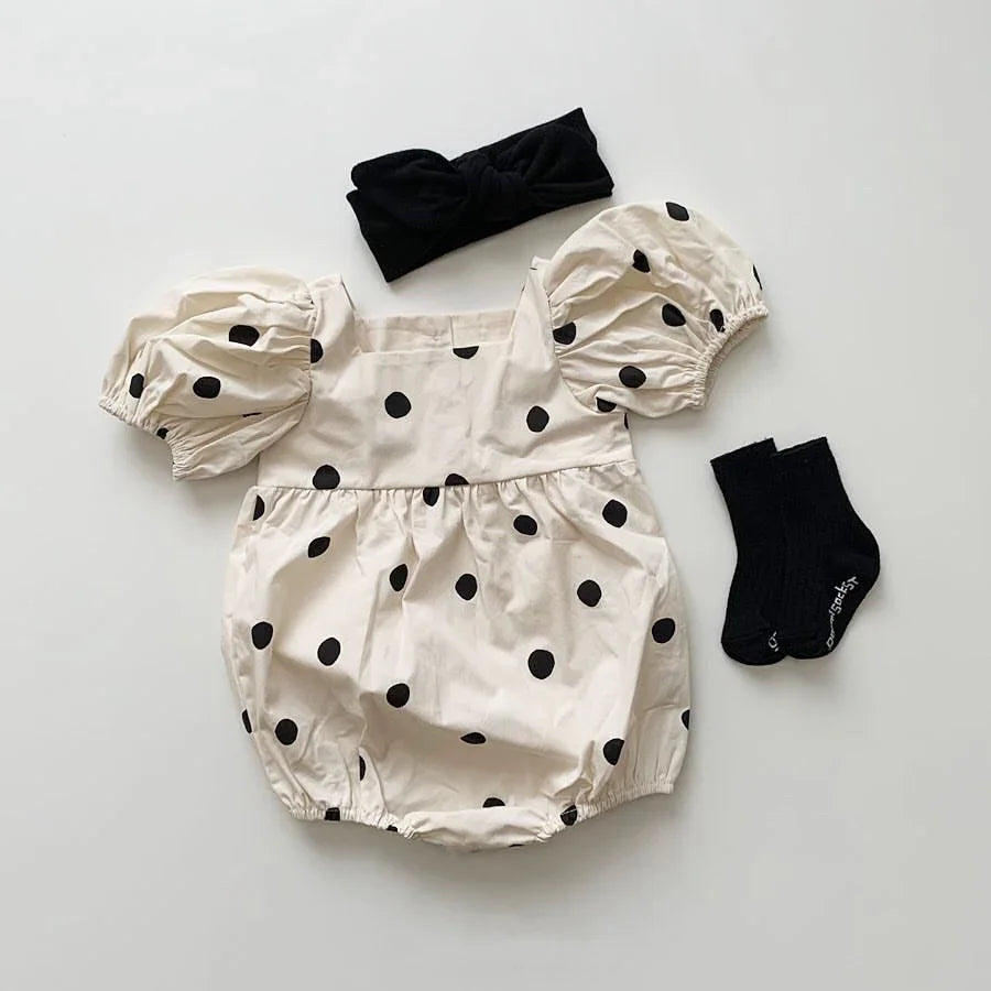 Cream baby romper with black polka dots, puff sleeves, black headband, and black socks laid flat