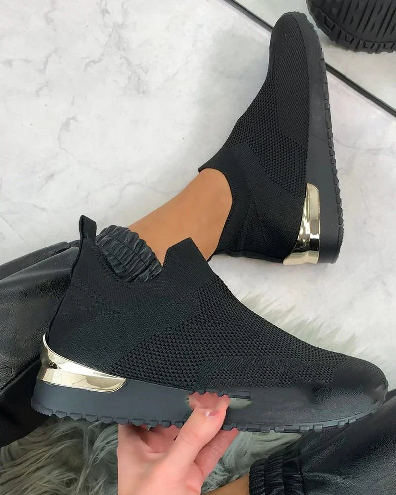 Black knit slip-on sneakers with gold metallic heel detail worn with black elastic cuff pants on marble floor