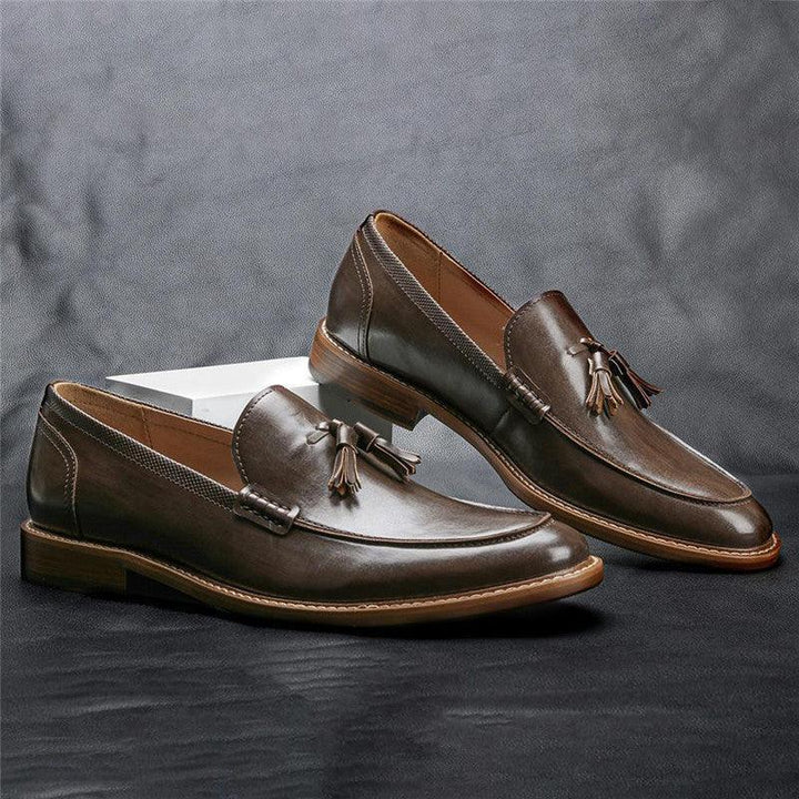 Pair of brown leather tassel loafers with tan soles displayed on dark surface against gray background