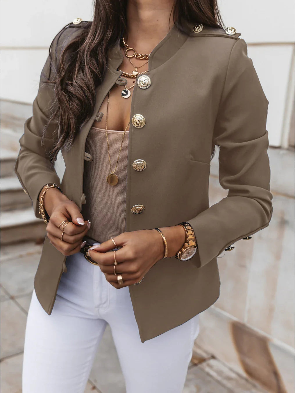 Woman wearing olive green military-style blazer with gold buttons, white pants, layered gold necklaces, and gold bracelets