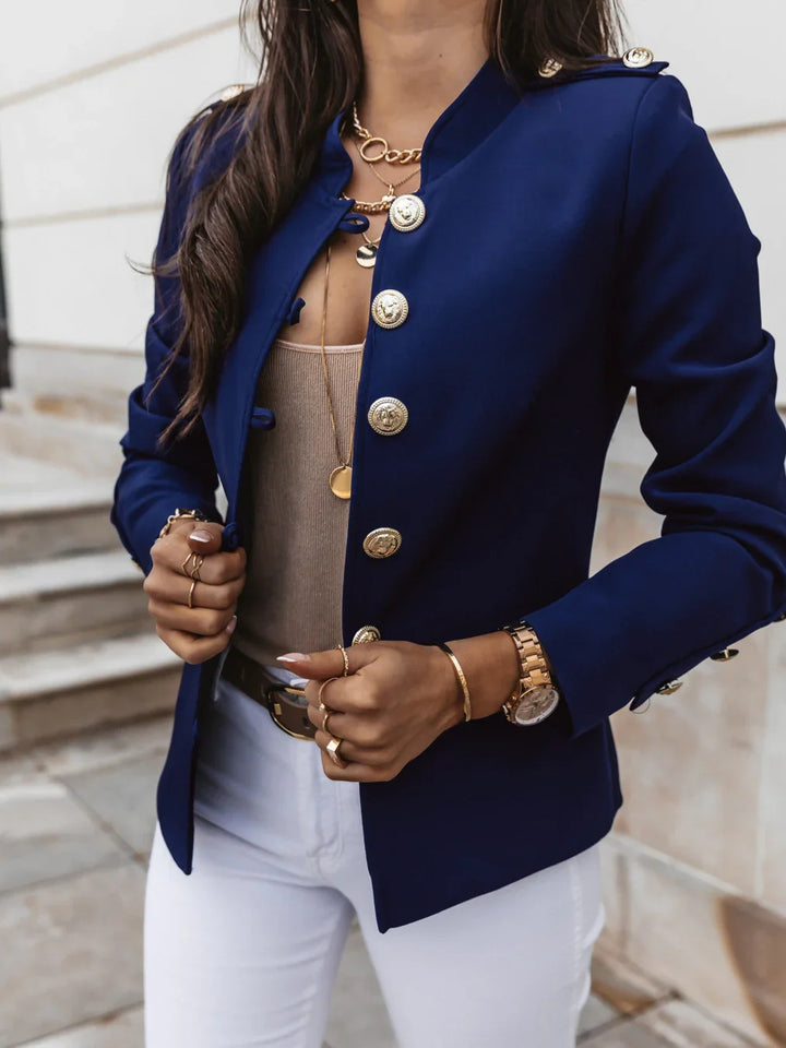 Woman wearing navy blue blazer with gold buttons, beige top, white pants, and gold jewelry accessories