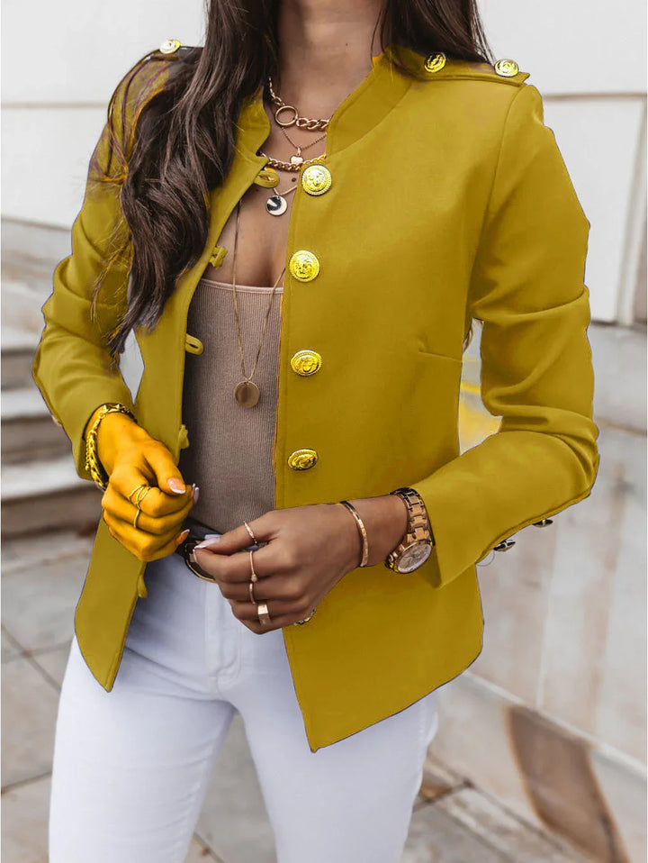 Woman wearing a mustard yellow military-style blazer with gold buttons, white pants, and layered necklaces