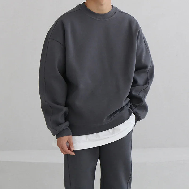 person wearing dark gray oversized sweatshirt and matching pants with white shirt layer, minimalist background