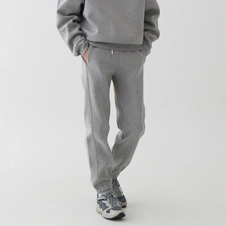 Person wearing gray sweatpants and matching sweatshirt with sneakers on a plain background