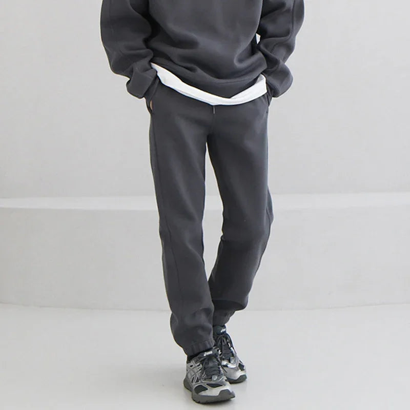 Person wearing dark gray jogger pants and matching sweatshirt with sneakers on a light background