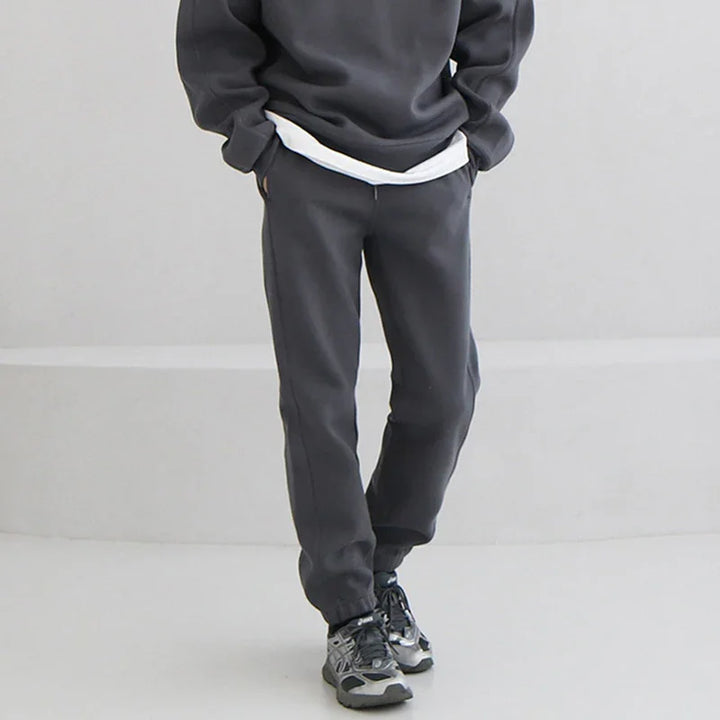 Person wearing dark gray jogger pants and matching sweatshirt with sneakers on a light background