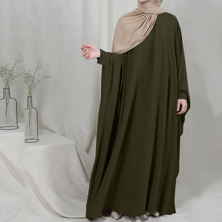 Woman wearing olive green abaya and beige hijab standing next to minimalist white cloth and vases with flowers