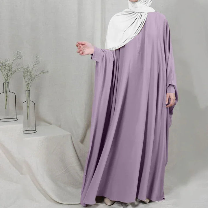 Woman wearing lavender modest abaya dress with white hijab in minimalist indoor setting with glass vases