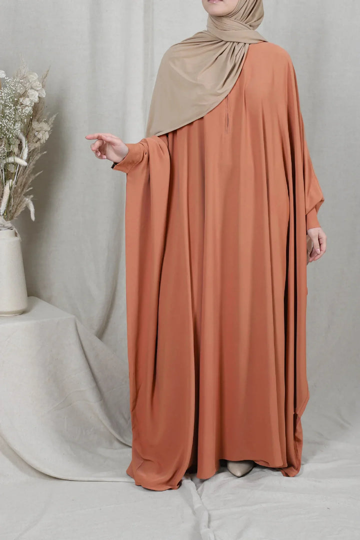 Woman wearing terracotta-colored long abaya dress with beige hijab, standing indoors with neutral backdrop
