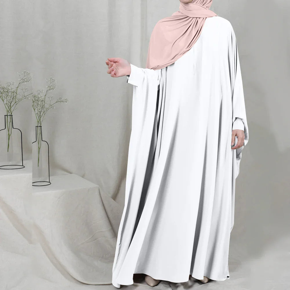 Woman wearing white abaya dress and light pink hijab standing indoors with decorative vases and fabric backdrop