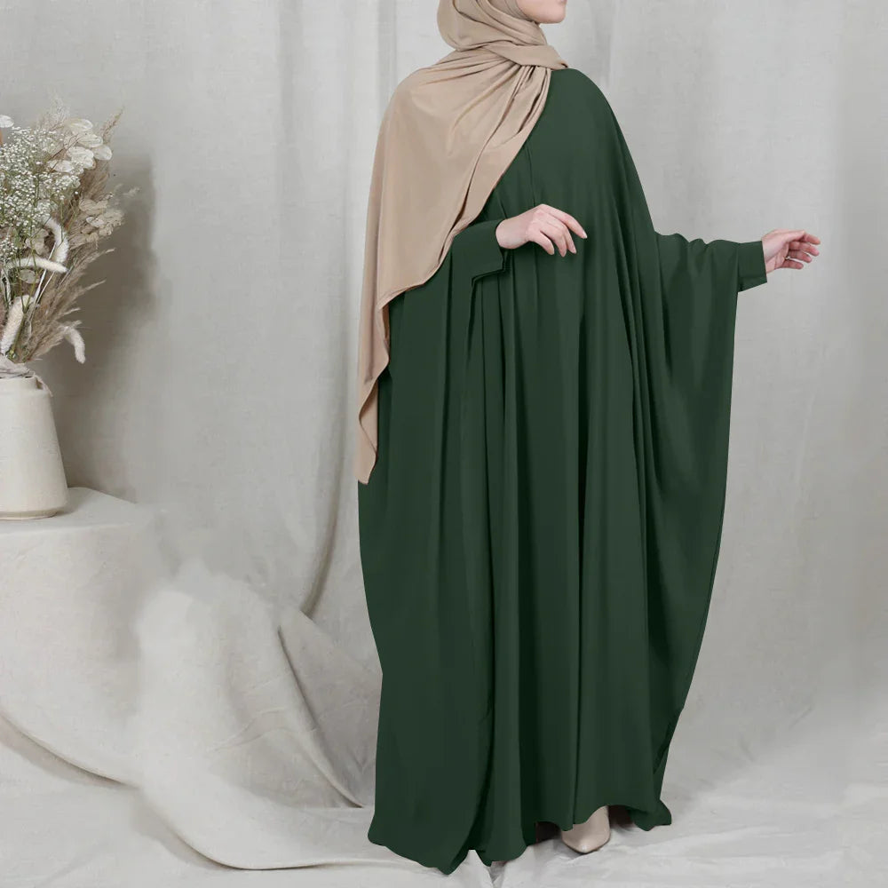Woman wearing a dark green abaya and beige hijab standing next to a vase with dried flowers