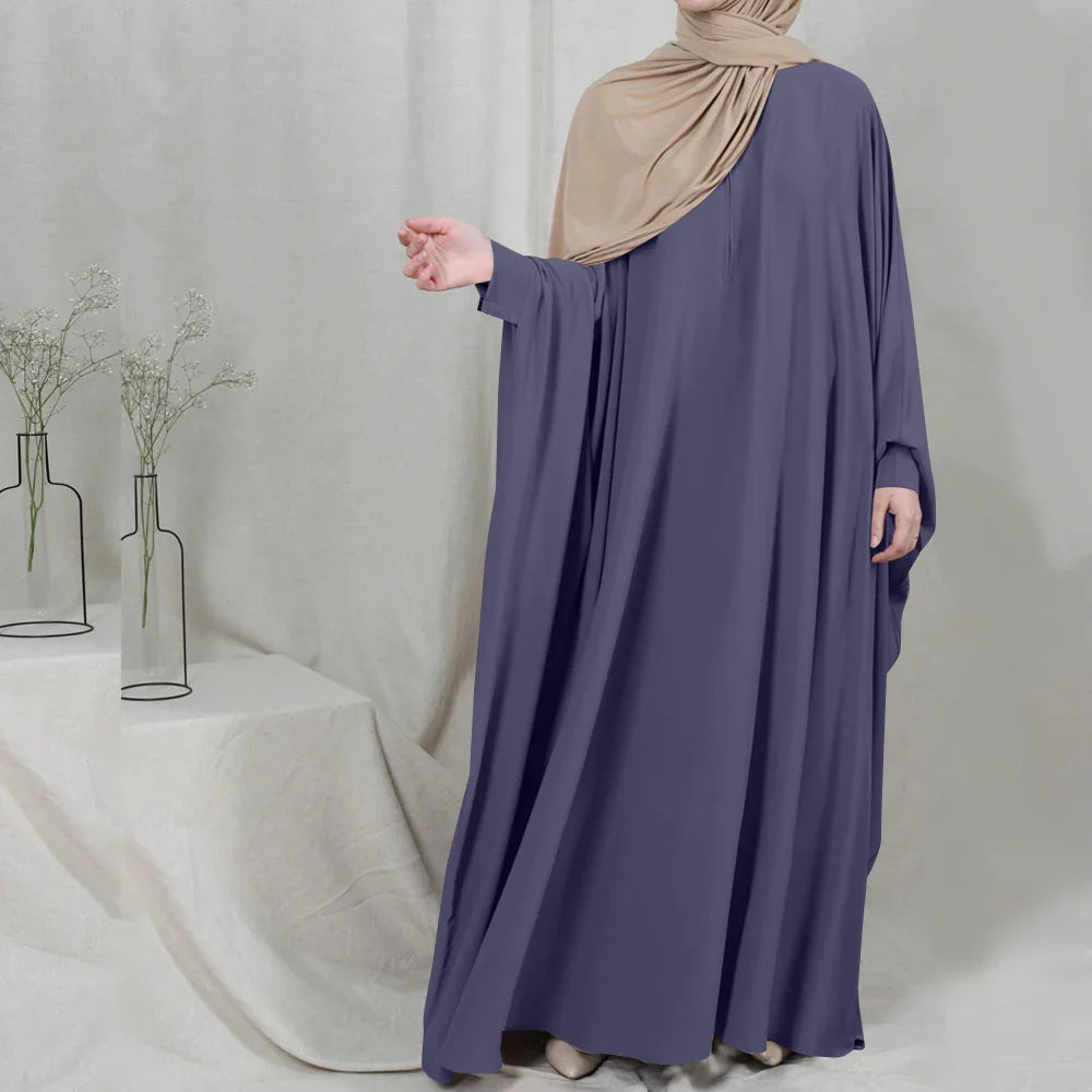 Woman wearing a loose purple abaya and beige hijab, standing against a neutral fabric backdrop with minimalistic vases and white flowers