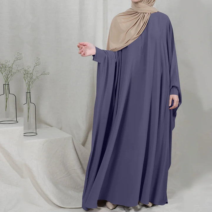 Woman wearing a loose purple abaya and beige hijab, standing against a neutral fabric backdrop with minimalistic vases and white flowers
