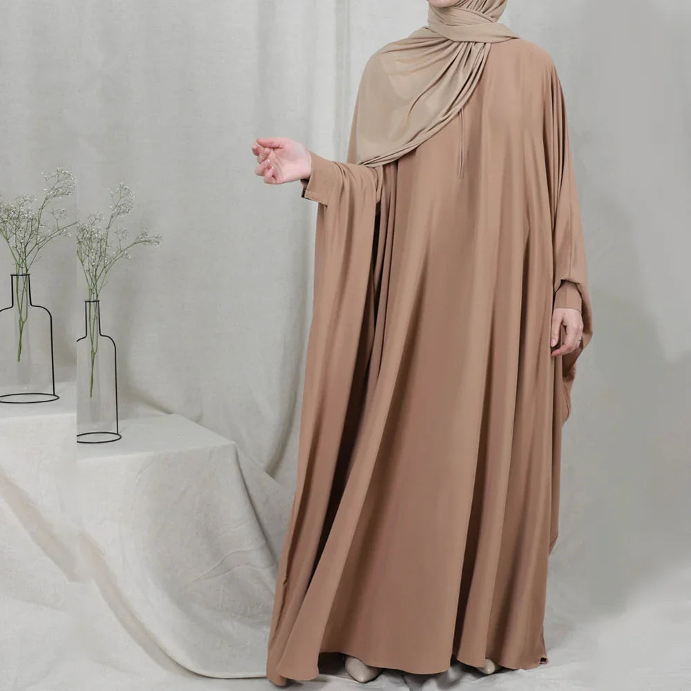 Woman wearing light brown modest abaya dress and matching hijab in minimalistic indoor setting with flower vases