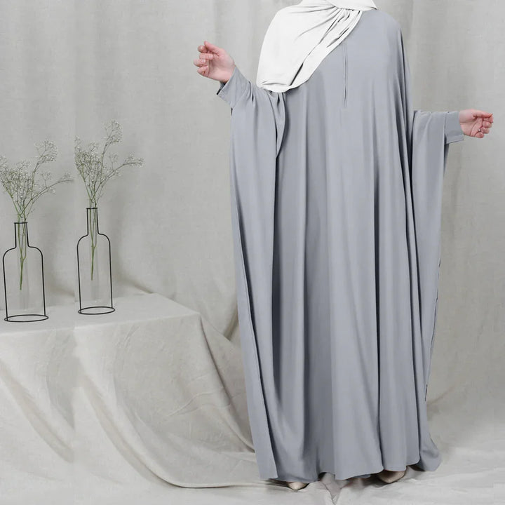 Woman wearing loose gray abaya and white hijab standing beside table with two minimalist vases