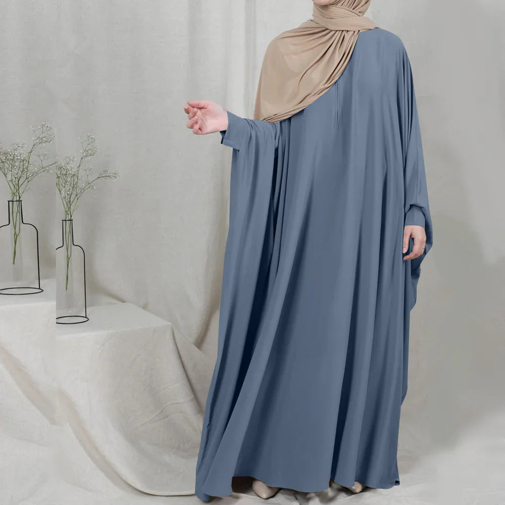Woman wearing a loose blue abaya and beige hijab standing indoors with minimalistic background