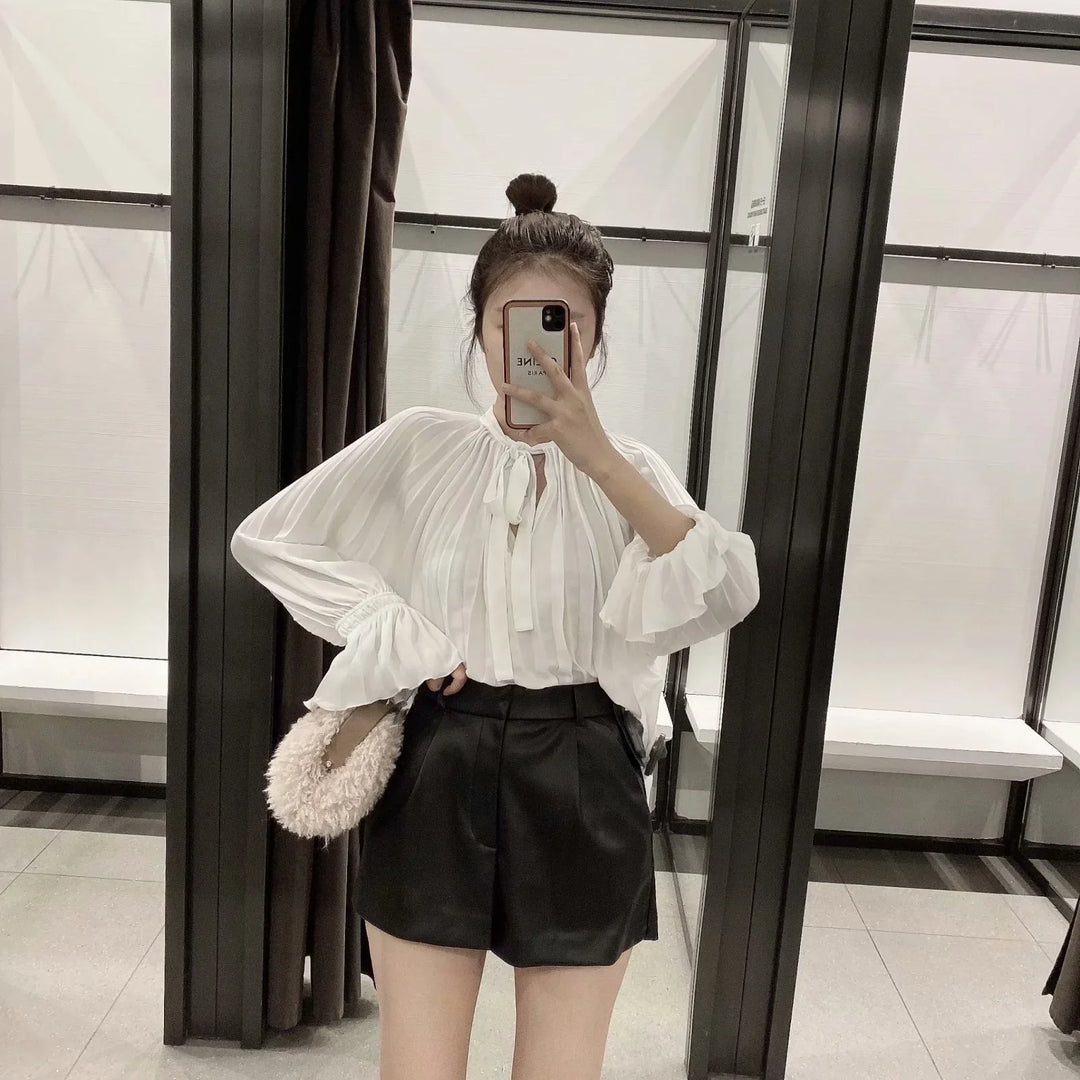 Woman taking mirror selfie wearing white pleated blouse and black high-waisted shorts, holding fluffy bag