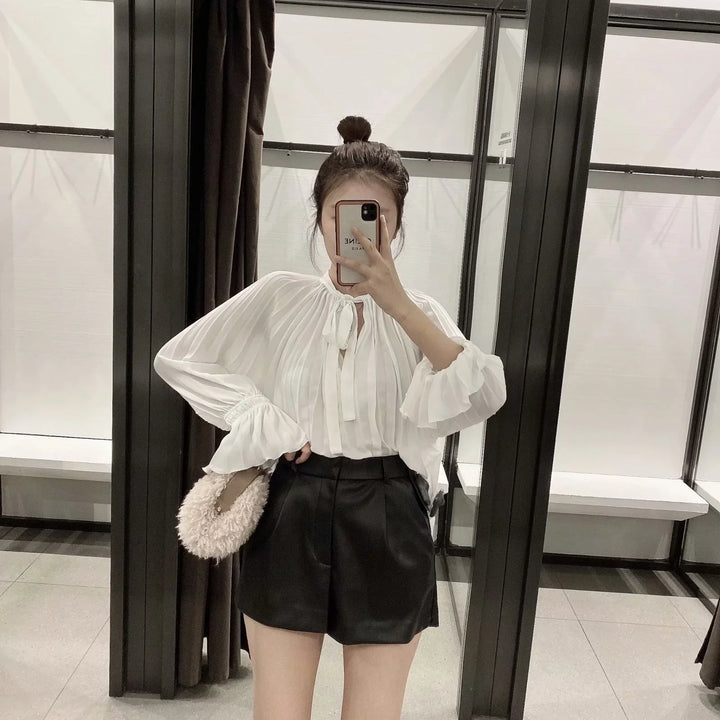 Woman taking mirror selfie wearing white pleated blouse and black high-waisted shorts, holding fluffy bag