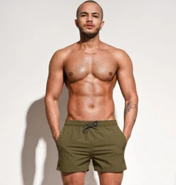 Fit muscular man wearing olive green swim trunks standing against a plain white background