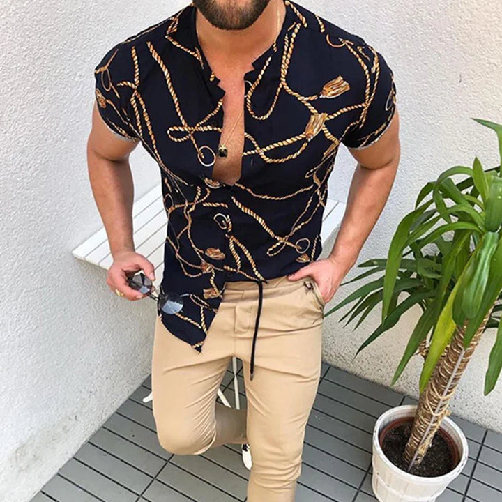Man wearing black rope print short-sleeve shirt and beige pants holding sunglasses near potted plant on patio