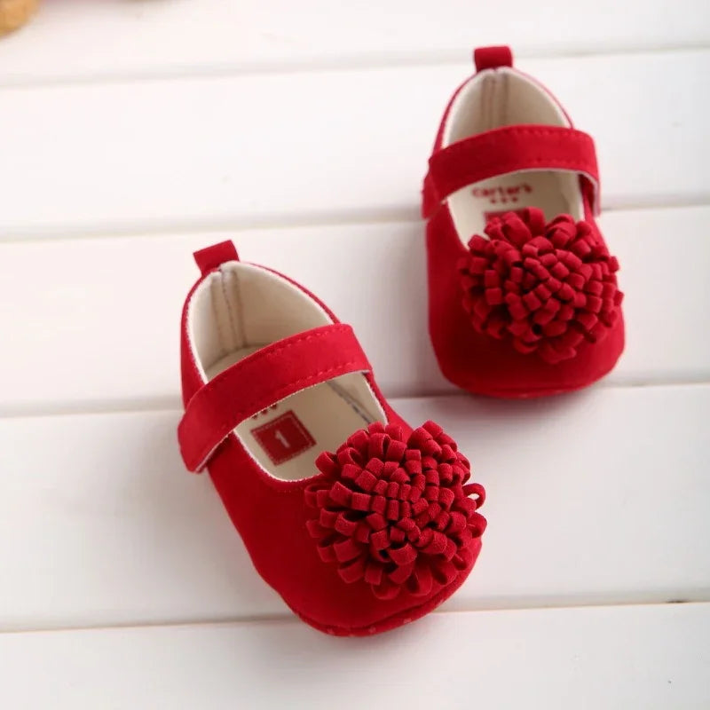 Pair of red baby shoes with large textured flower decorations and velcro straps on white wooden background