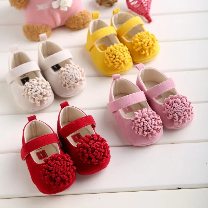 Four pairs of colorful baby shoes in red, pink, yellow, and white with flower decorations on a white wooden surface