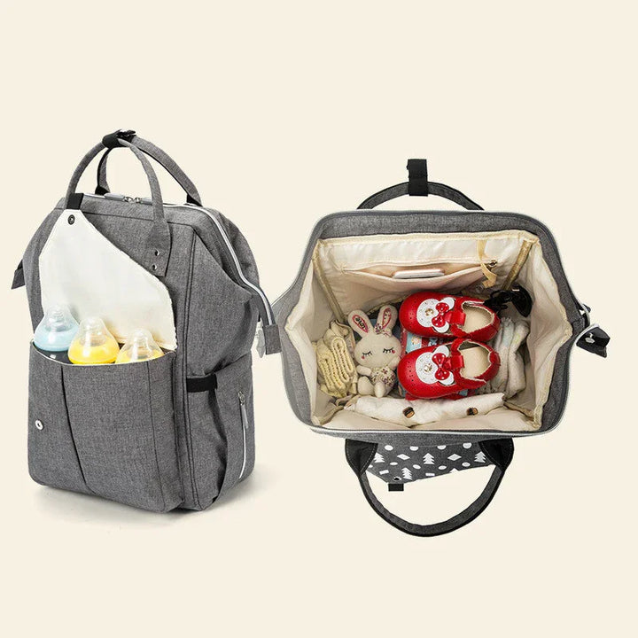 Gray baby diaper backpack with multiple compartments holding baby bottles, plush toy, red baby shoes, and clothes