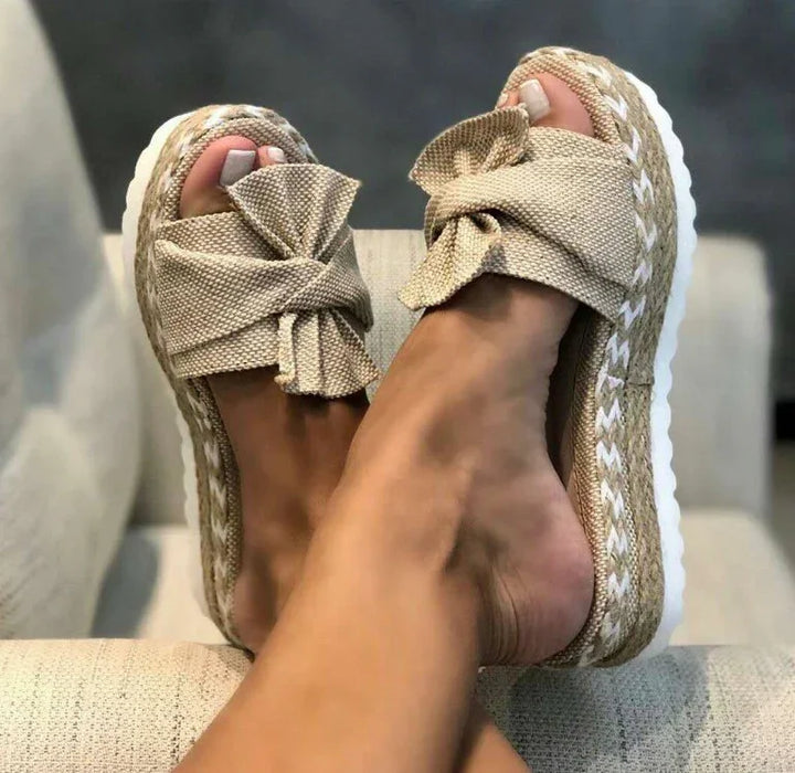 Close-up of feet wearing beige platform sandals with large fabric bows on top, resting on a light-colored textured surface