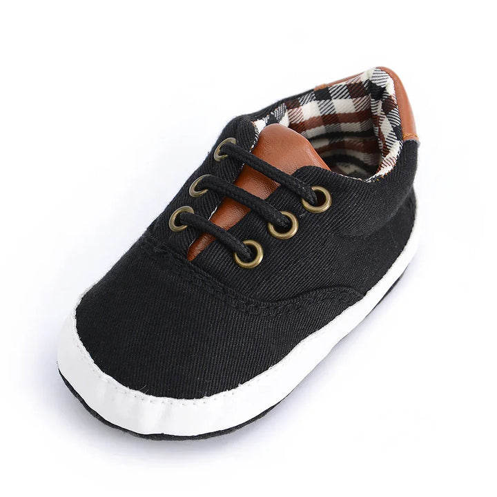 Black baby shoe with white sole, brown faux leather tongue, and plaid inner lining