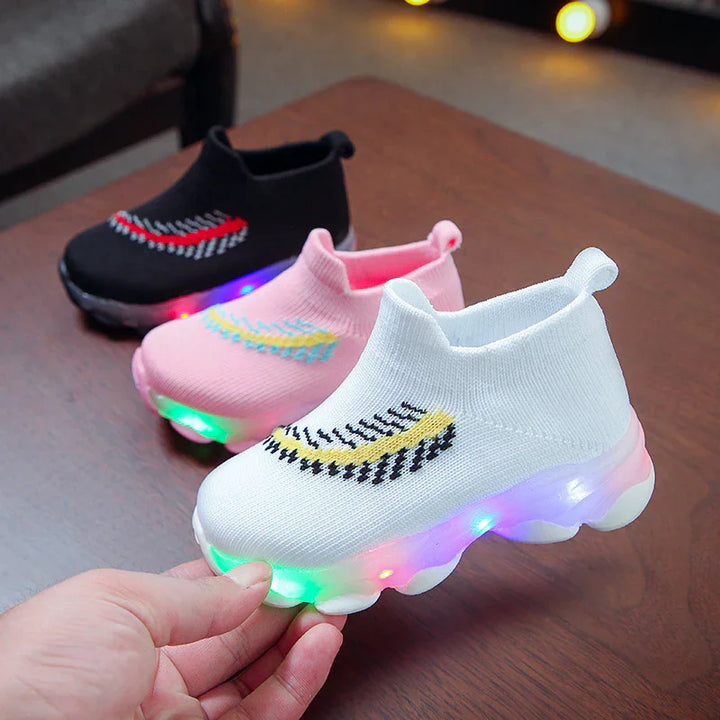 Three pairs of kids light-up sneakers in white, pink, and black with colorful soles on a wooden table