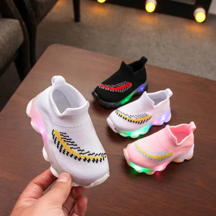 Hand holding white kids light-up sneakers with three other pairs on brown table in black, white, and pink colors