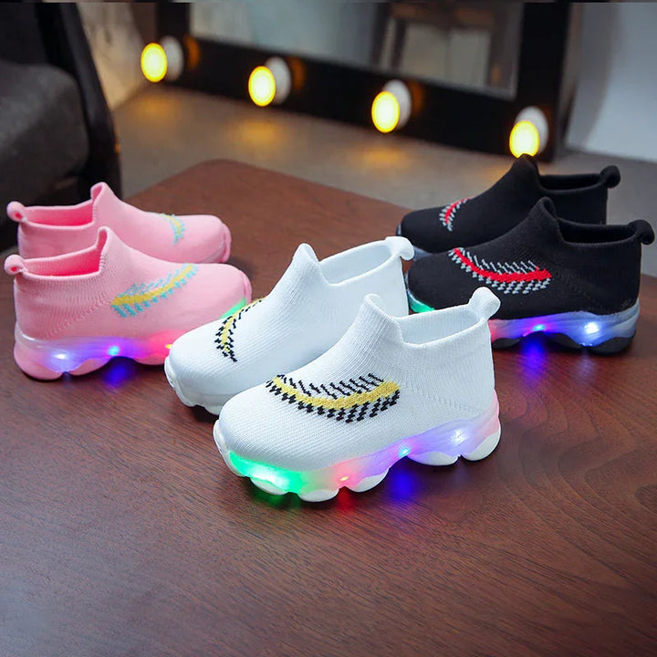 Three pairs of kids light-up sneakers in pink, white, and black on wooden floor with glowing multicolor LED soles