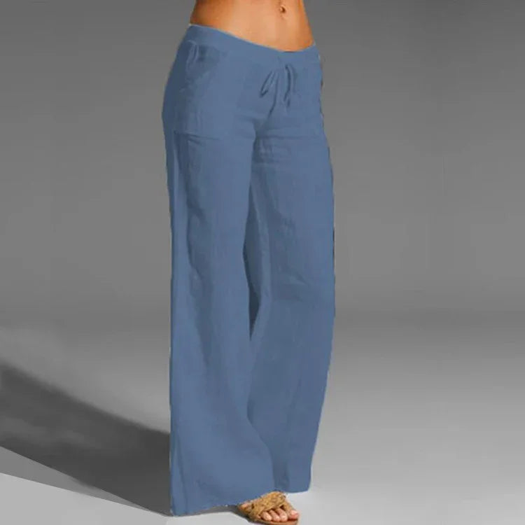 Model wearing high-waisted loose blue linen pants with drawstring and sandals on gray background
