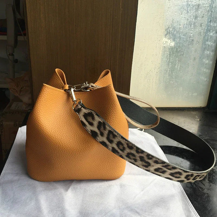 Tan leather bucket bag with leopard print strap on white cloth near foggy window