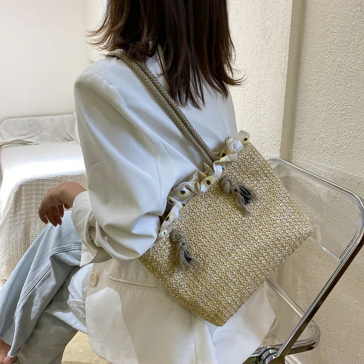 Woman wearing white blazer sitting on transparent chair with woven straw bag featuring tassels and ruffled edge