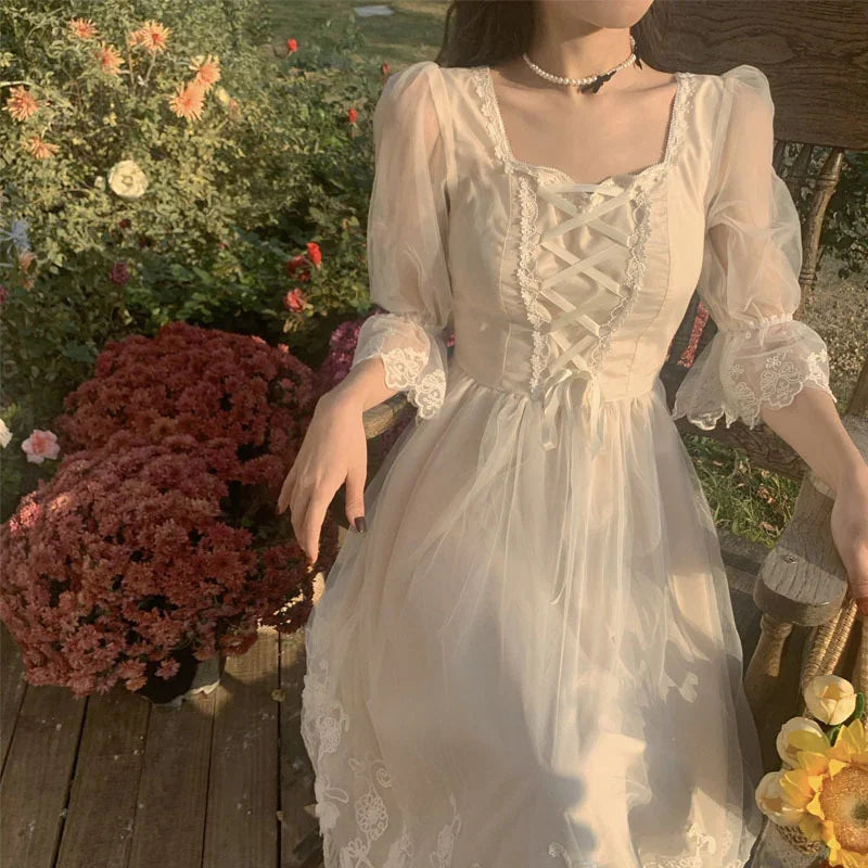 Woman in vintage lace cream dress sitting on wooden chair beside pink flowers in garden