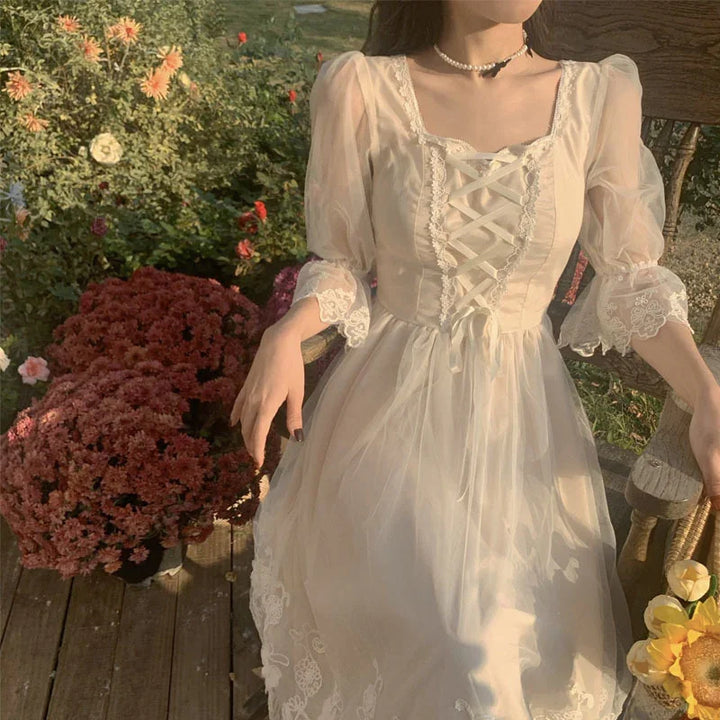 Woman in vintage lace cream dress sitting on wooden chair beside pink flowers in garden