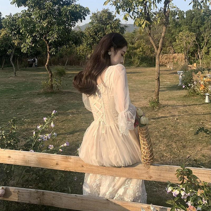 Woman in vintage lace dress sitting on wooden fence in sunlit garden holding woven bag with flowers
