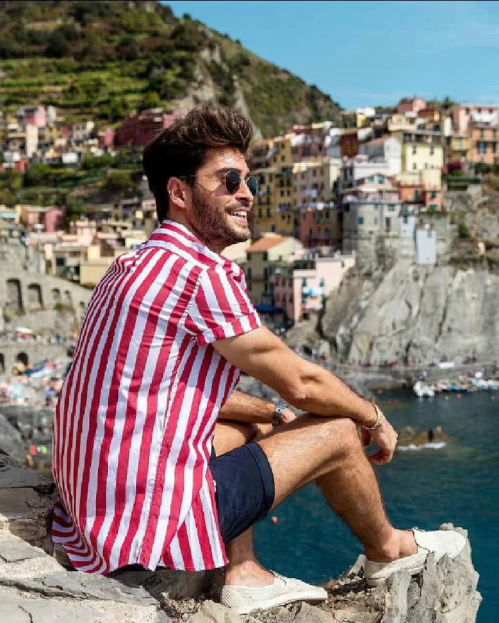 Smiling man in red and white striped shirt and sunglasses sitting on cliff overlooking colorful coastal village and sea
