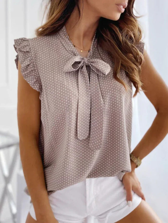 Woman wearing a taupe sleeveless polka dot blouse with ruffle sleeves and bow tie neckline paired with white shorts