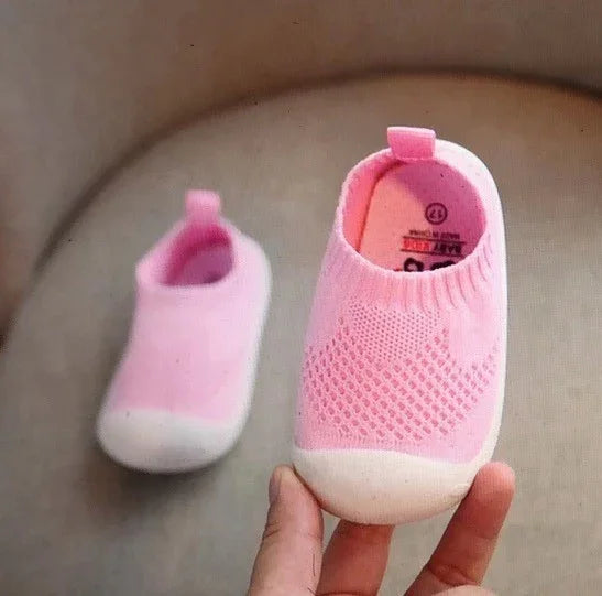 Soft pink baby shoes with white soles held by hand on beige background