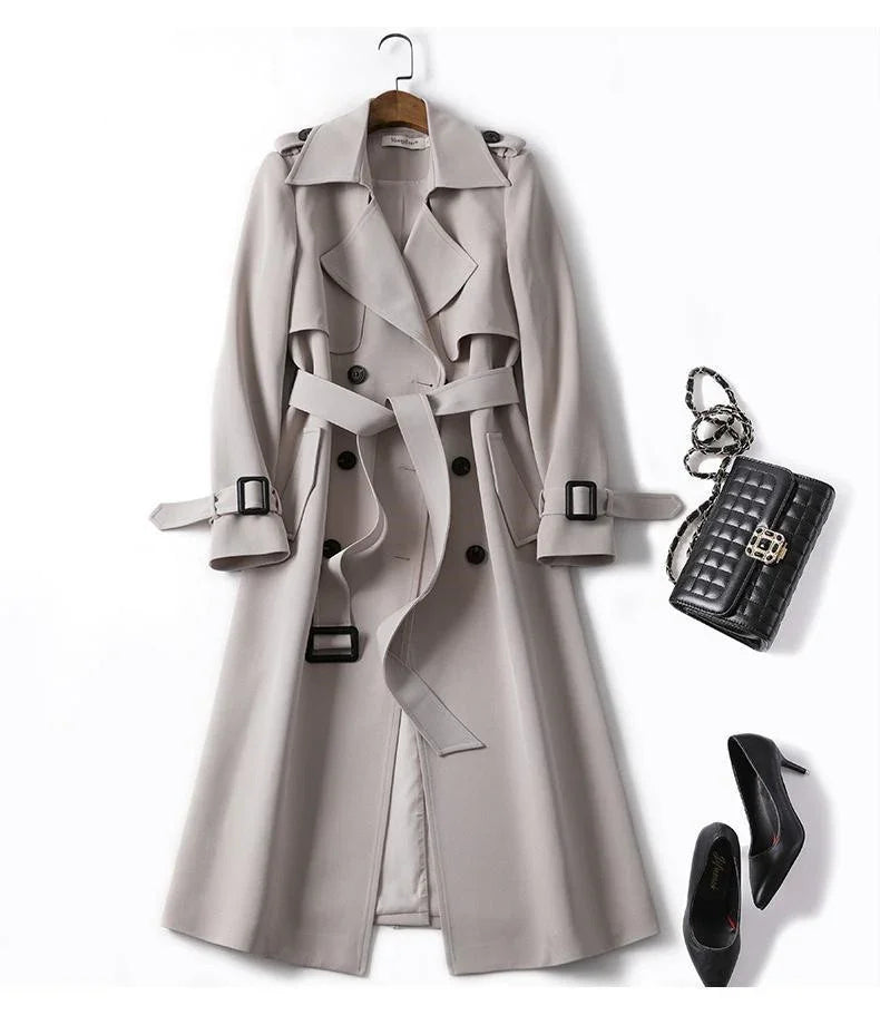 Beige long trench coat with belt, black quilted handbag, and black high heel shoes on white background