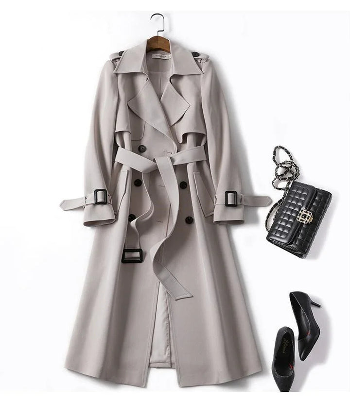 Beige long trench coat with belt, black quilted handbag, and black high heel shoes on white background