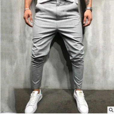 Man wearing fitted grey tapered jogger pants with front seams and white sneakers against a grey wall