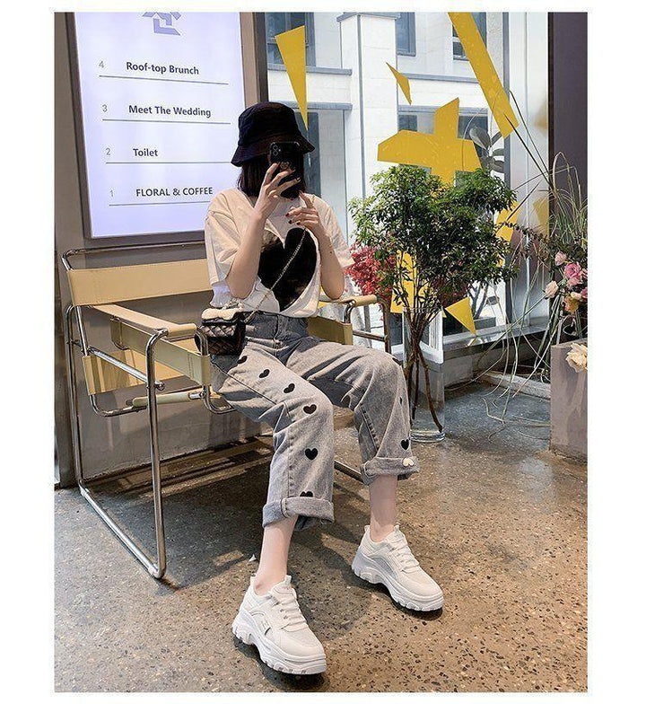 Person wearing heart print jeans, white sneakers, black bucket hat, sitting indoors with plants and art