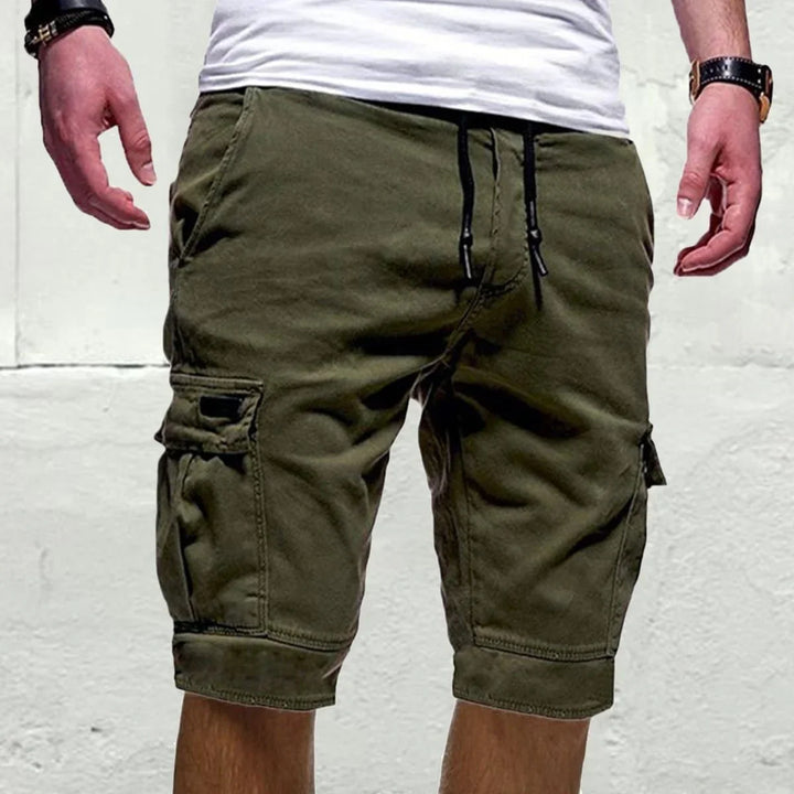 Man wearing olive green cargo shorts with front pockets and black drawstring against white wall