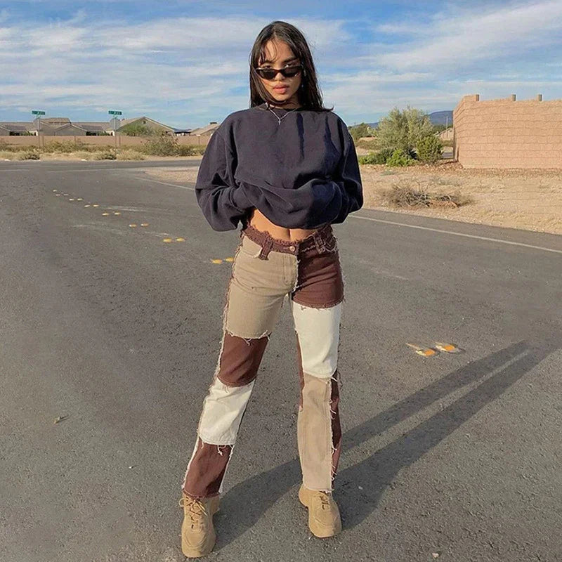 Young woman in black cropped sweatshirt, patchwork jeans, sunglasses, and beige boots standing on empty road