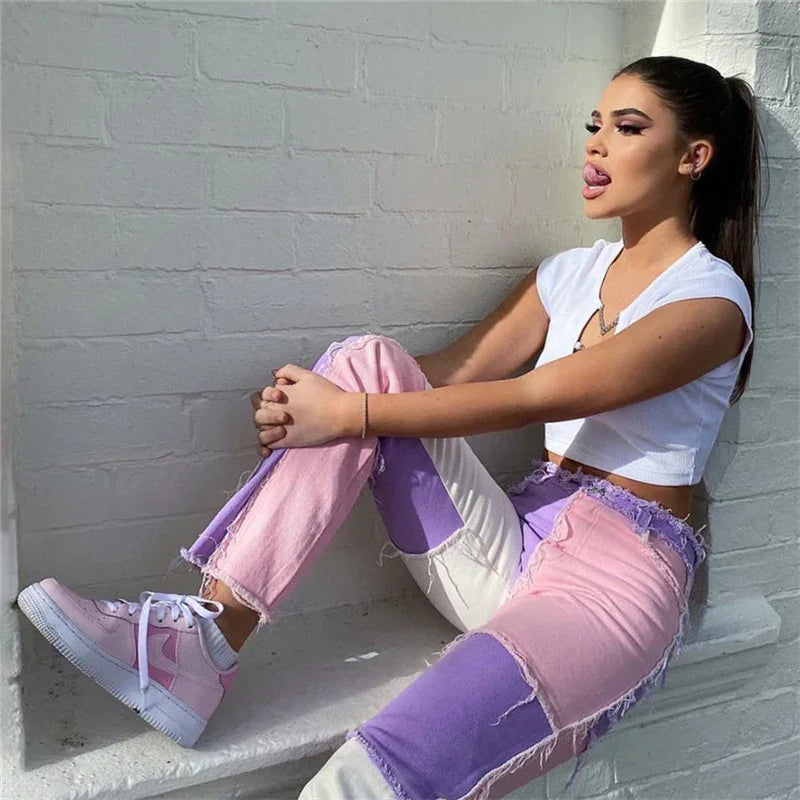 Young woman in colorblock pink and purple jeans and white crop top sitting against white brick wall in sunlight