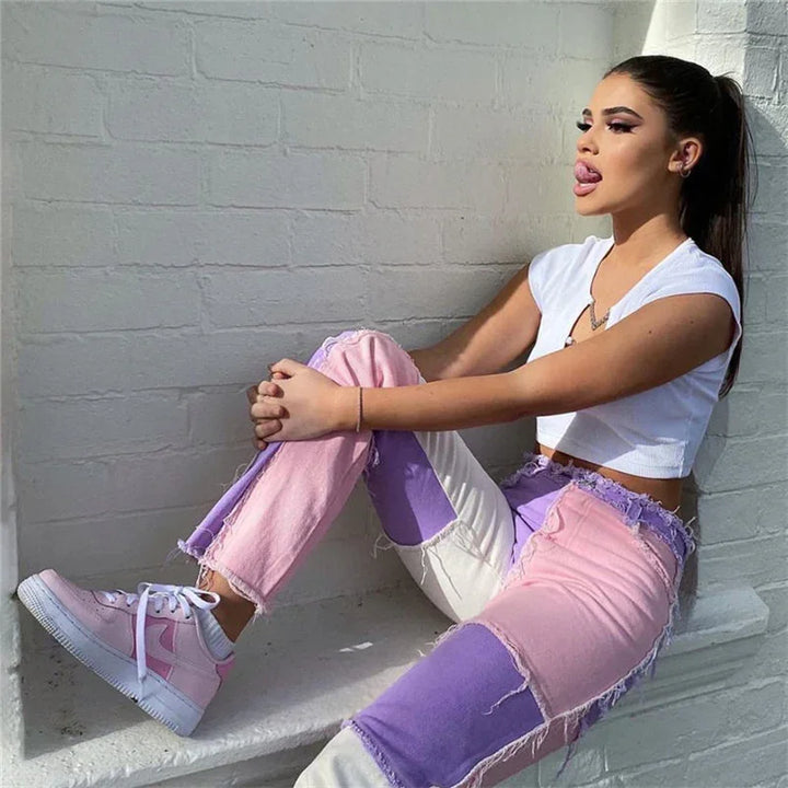 Young woman in colorblock pink and purple jeans and white crop top sitting against white brick wall in sunlight